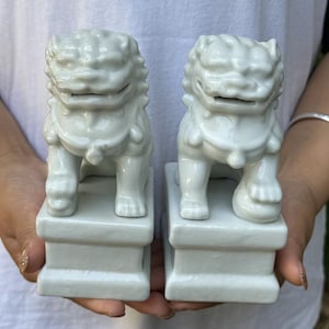 May include: A pair of matching, off-white ceramic Foo Dog statues. Each statue is in a seated position on a rectangular base. The Foo Dogs have detailed features, including a mane, facial features, and a decorative collar.