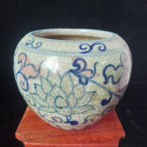 May include: A small, round ceramic pot with a cream-colored base and intricate blue and pink floral patterns. The pot has a cracked glaze effect and sits on a small, reddish-brown wooden stand. The design is reminiscent of traditional Asian pottery.