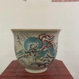 May include: A ceramic teacup with a detailed dragon design. The dragon is depicted in shades of blue, red, and yellow against a gray background. The cup has a rounded shape and sits on a wooden stand.