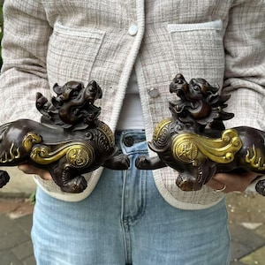 May include: Two bronze-colored mythical creature statues with gold accents. The statues have detailed features, including open mouths and ornate designs on their bodies. The statues are held up in front of a person wearing a light-colored jacket and blue jeans.