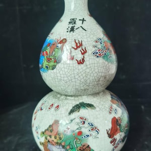 May include: A white, double-gourd-shaped ceramic vase with a crackle glaze. The vase features colorful illustrations of figures and Chinese characters. The base is a dark wood color.