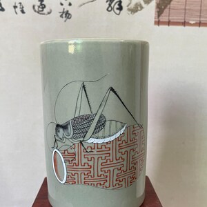 May include: A cylindrical, celadon-colored ceramic vase featuring a detailed illustration of a grasshopper. The grasshopper is depicted in black and gray, resting on a red and white patterned design. The vase is displayed on a small, dark wooden stand.