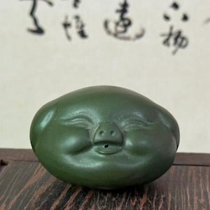 May include: A dark green ceramic sculpture of a smiling pig with closed eyes and a small snout. The sculpture is smooth and rounded, with a matte finish. The background features blurred calligraphy.
