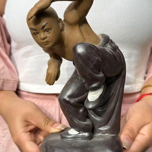 May include: A matte brown ceramic figurine of a person in a crouching martial arts pose. The figure has a shaved head, dark eyebrows, and is wearing dark brown loose-fitting trousers with white accents on the shoes, set on a solid base.