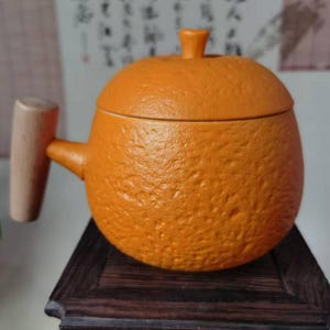 May include: A ceramic teapot with a lid, handle, and spout, designed to resemble an orange. The teapot is a vibrant orange color with a textured surface, and a light-colored wooden handle. The teapot is sitting on a dark wooden surface.