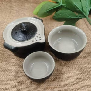 May include: A ceramic tea set with a black and gray color scheme. The set includes a teapot with a lid and two matching teacups. The teapot has a unique shape with a spout and handle. The teacups are small and round.