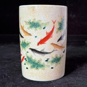 May include: A cylindrical ceramic cup with a cream-colored, crackled finish. The cup is decorated with colorful koi fish in shades of red, orange, black, and gray, swimming among green aquatic plants. A unique piece for home decor.