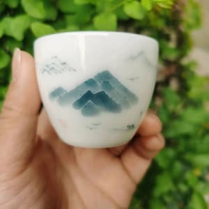 May include: A small, white ceramic teacup with a hand-painted mountain landscape in shades of blue and gray. The design features stylized mountains and a few small birds. The cup has a smooth, glossy finish.