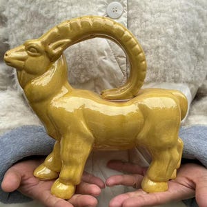 May include: A ceramic goat figurine in a glossy yellow glaze. The goat has large curved horns and is standing on four legs. The figurine is held in someone's hands.