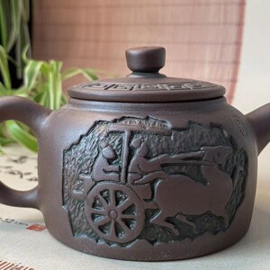 May include: Brown ceramic teapot with a lid and handle. The teapot features a detailed relief carving of a horse-drawn cart with figures. The lid has a decorative pattern. A spout and a curved handle complete the design.