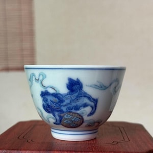 May include: White ceramic teacup with a blue and white illustration of a mythical creature. The cup has a rounded shape and a detailed design, with blue accents and a decorative rim. The cup is sitting on a wooden surface.