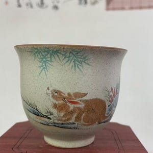 May include: A ceramic teacup with a hand-painted design of a brown rabbit, bamboo, and flowers. The cup has a speckled, off-white background and a brown rim. The rabbit is looking up, and the cup sits on a wooden stand.