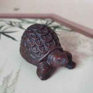 May include: A small, intricately carved wooden turtle figurine. The dark brown turtle has a detailed shell pattern and visible legs and head. The figurine is displayed on a light-coloured surface.