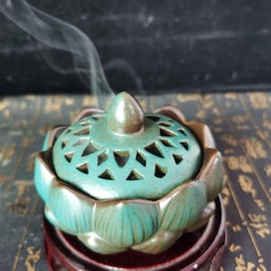 May include: A teal ceramic incense burner with a lotus flower design. The lid has geometric cutouts and a brown finial. Wisps of smoke rise from the burner, suggesting use. The burner sits on a dark wooden base.