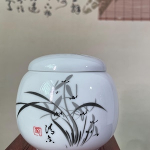 May include: A white ceramic jar with a lid, featuring a hand-painted design of black and gray bamboo stalks and leaves. The jar has a red square stamp and black calligraphy. The jar is sitting on a dark wooden surface.