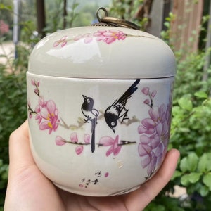 May include: A white ceramic jar with a lid, featuring a floral and bird design. The jar has a rounded shape and a small metal loop on top. The design includes pink flowers and black and white birds. The jar is held in a hand.