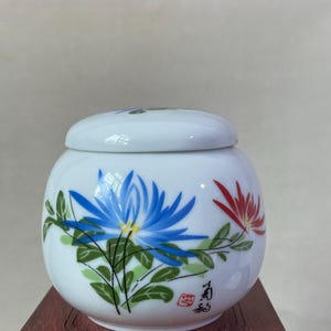 May include: A white ceramic jar with a lid, adorned with hand-painted blue and red floral designs. Green leaves and stems complement the flowers. The jar sits on a dark wooden stand, showcasing the detailed artwork.