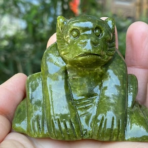 May include: A carved green jade monkey figurine. The monkey is sitting with its legs crossed and its hands resting on its knees. The figurine has detailed facial features and a smooth, polished surface. The monkey is a symbol of good luck and fortune.