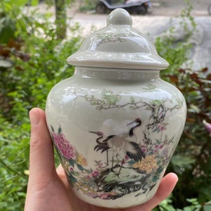 May include: A pale green ceramic jar with a lid, featuring a painted design of two cranes, flowers, and foliage. The jar has a rounded body and a small knob on the lid. The artwork includes pink, yellow, and black accents.