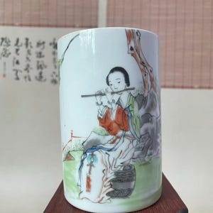 May include: A cylindrical white porcelain brush pot featuring a hand-painted illustration of a figure in traditional orange and green robes playing a flute while seated on a dark gray stool, accented by black ink calligraphy and a tree branch.