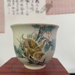 May include: A ceramic teacup with a beige background and a painted monkey design. The monkey is brown and tan, with a red face, and is climbing on a tree branch with green and blue leaves. The cup is on a red wooden surface.