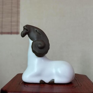 May include: A ceramic ram figurine with a white body and a dark brown head and horns. The ram is in a seated position on a wooden surface. The figurine has a smooth, glossy finish and is likely a decorative item.