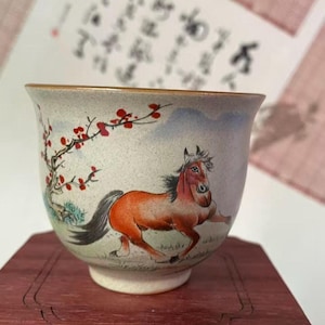 May include: A ceramic teacup with a hand-painted design of a running horse in shades of orange and brown. The cup also features a flowering tree with red blossoms. The rim is a warm brown color, and the cup sits on a white base.