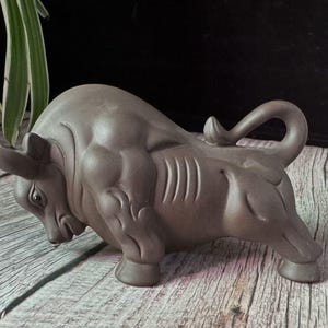 May include: A gray ceramic bull figurine in a charging pose. The sculpture has detailed musculature and curved horns. The bull's tail is curled upwards. The figurine is on a wooden surface.