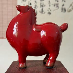 May include: A glossy, red ceramic horse figurine. The sculpture has a stylized form with a rounded body, short legs, and a small tail. The horse is standing on a dark wooden base. The background features a blurred wall with calligraphy.