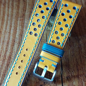 Può includere: Cinturino per orologio in pelle giallo senape con dettagli blu. Il cinturino presenta cuciture blu e perforazioni circolari. È presente una fibbia argentata. Il cinturino è esposto su una superficie di legno.