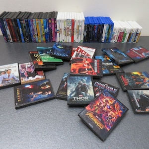 Peut inclure: Une collection de mini-DVD et Blu-ray de films. L'image pr&eacute;sente des piles de bo&icirc;tiers de diff&eacute;rentes couleurs, avec plusieurs couvertures de films individuelles &eacute;parpill&eacute;es devant. Les titres incluent "Avatar", "Thor" et "The Avengers".