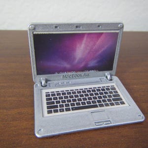 May include: A miniature silver MacBook Air laptop toy with a screen displaying a purple and pink image. The keyboard has black keys and white lettering. The laptop is open and sits on a brown surface.