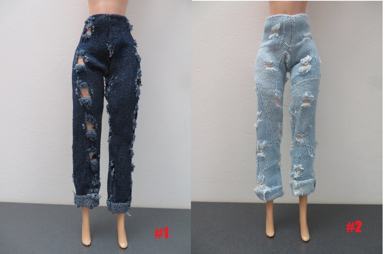 May include: Two pairs of doll jeans: one dark blue, the other light blue. Both have distressed details with ripped sections. The dark blue pair has rolled-up cuffs, and the light blue pair also has rolled cuffs. The jeans are displayed on doll legs.