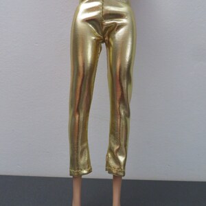 May include: Gold metallic pants with a slight flare at the ankle. The pants are made of a shiny, reflective material and have a fitted waist. The pants are designed for a doll.