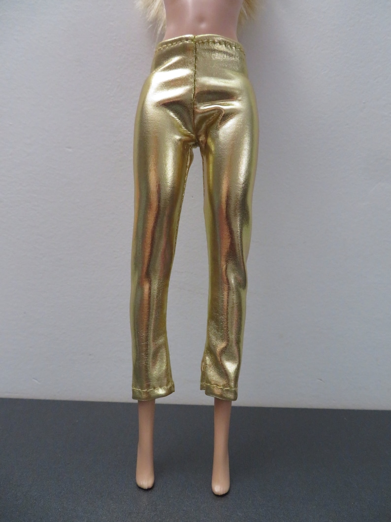 May include: Gold metallic pants with a slight flare at the ankle. The pants are made of a shiny, reflective material and have a fitted waist. The pants are designed for a doll.