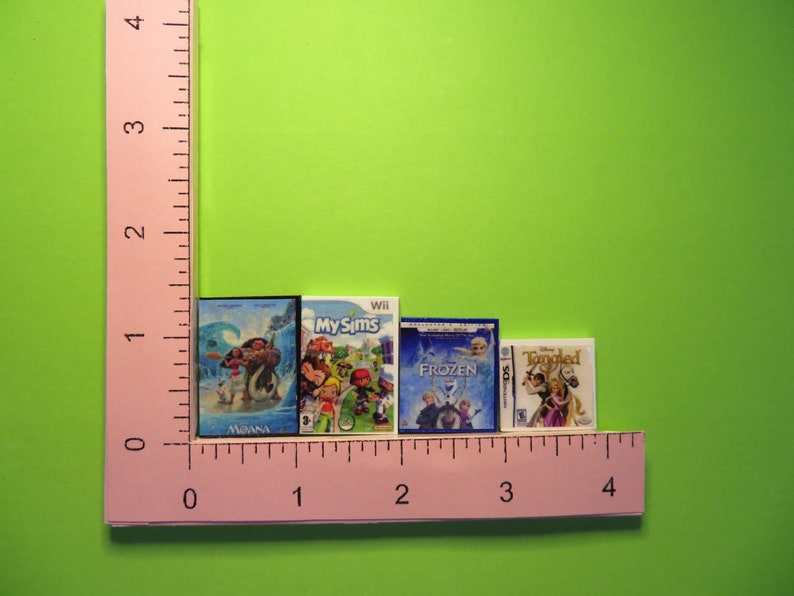 Peut inclure: Miniatures de jeux vid&eacute;o pour Wii et Nintendo DS, dont "Moana", "My Sims", "Frozen" et "Tangled". Les bo&icirc;tes sont dispos&eacute;es en rang&eacute;e, avec une r&egrave;gle rose en arri&egrave;re-plan. Les bo&icirc;tes mesurent environ 2.5 cm de hauteur.