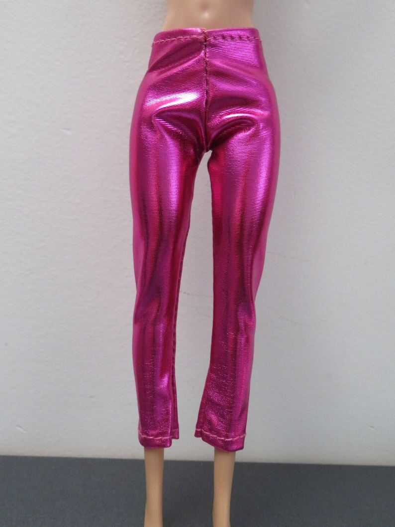 May include: Bright pink, metallic-finish pants. The pants have a high waist and a straight leg cut, with a slight flare at the ankle. The fabric has a textured appearance, reflecting light. The pants are designed for a doll.