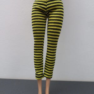 May include: Black and yellow striped leggings. The leggings are made of a knit fabric and feature horizontal stripes. The doll-sized pants have a fitted design and are displayed on a doll.