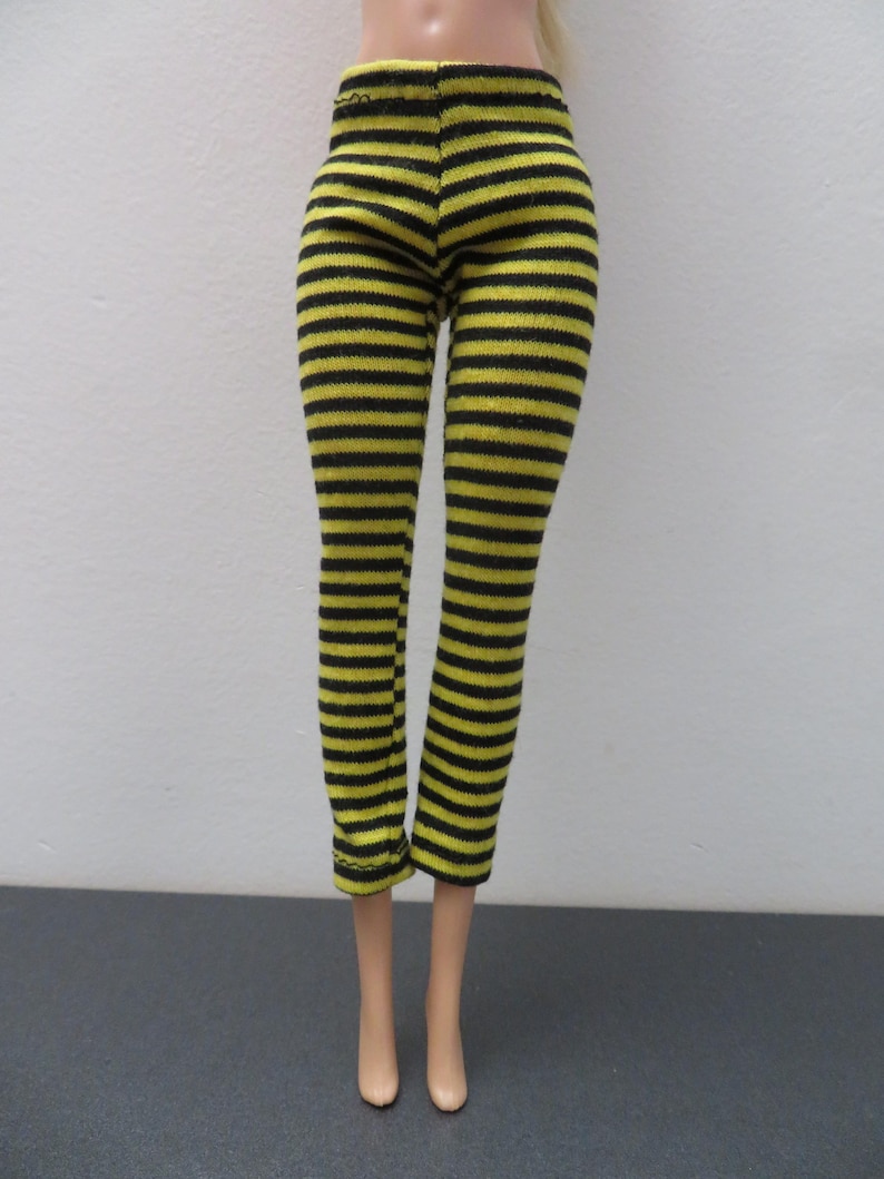 May include: Black and yellow striped leggings. The leggings are made of a knit fabric and feature horizontal stripes. The doll-sized pants have a fitted design and are displayed on a doll.
