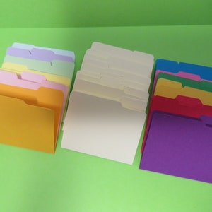May include: A collection of colorful file folders in shades of yellow, pink, purple, blue, green, and cream. Each folder has a tab for labeling. The folders are arranged on a bright green surface.