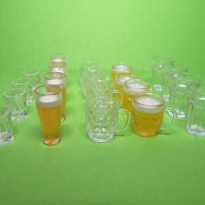 May include: A collection of miniature beer glasses and shot glasses arranged on a green surface. The beer glasses are filled with a yellow liquid and topped with white foam. The shot glasses are clear.