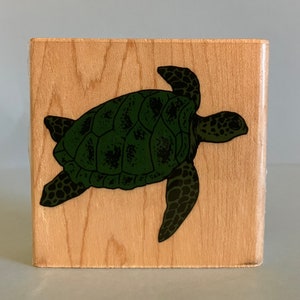 May include: A wooden block with a green sea turtle illustration. The turtle is swimming with its head and flippers visible. The block is made of light wood with a natural grain.