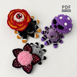 May include: Three crocheted plush creatures including a black spider with a red and orange rose-like top, a purple spider with a spotted mushroom cap, and a grey spider with a pink flower-like head. A PDF PATTERN label is visible in the corner.