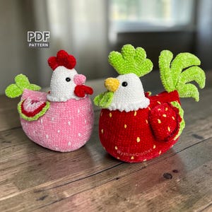 May include: Two handmade crochet hen figurines. One is pink with a strawberry design, the other is red with yellow seed details. Both have white heads, red combs, and green tail feathers. The text "PDF PATTERN" is in the upper left corner.