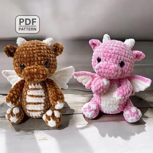 May include: Two crocheted dragon plush toys. One is brown with white details and wings, the other is pink with white details and wings. Both have black eyes and are seated. The image includes the text "PDF PATTERN".