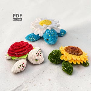 May include: Three crocheted turtle toys with flower-themed shells. One has a white daisy, another a red rose, and the third a yellow sunflower. The turtles are on a sandy surface. The text "PDF PATTERN" is in the upper left corner.