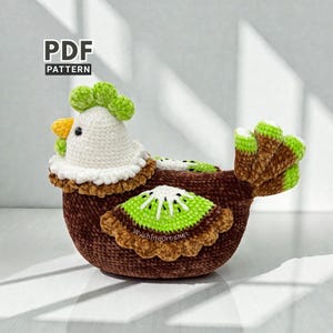 May include: A handmade crochet chicken-shaped basket in brown, white, yellow, and green. The chicken has a white head, a yellow beak, and a green comb. A kiwi slice is appliqued on the side. The text "PDF PATTERN" is in the upper left corner.