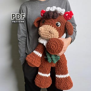 May include: A large, handmade crochet cow plushie in shades of brown, beige, white, red, and green. The plush toy has a detailed design with a white and beige muzzle, red and white accents on the ears, and a green leaf detail. The text "PDF PATTERN" is visible.