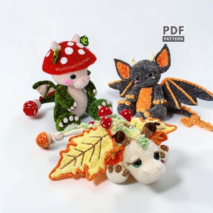 Peut inclure: Trois figurines de dragons au crochet. L'une présente un chapeau de champignon rouge à pois blancs, un corps et des ailes verts. Une autre est gris foncé avec des accents et des ailes orange. La troisième est crème avec de grandes ailes en forme de feuille jaune et de petits détails de champignons rouges. Le texte PDF PATTERN est visible.