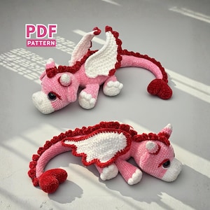 May include: Two handmade crochet dragons in pink, red, and white. The dragons have wings, horns, and a tail. The text "PDF PATTERN" is in the upper left corner. The dragons are lying on a light grey surface.
